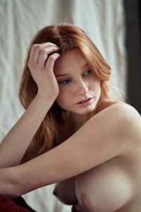 Woman with long auburn hair resting her hand against her head, bare shoulders, soft window light, pale drapery and red fabric behind.