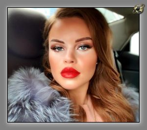 Woman with auburn waves and red lipstick in grey fur stole, inside car.