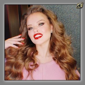 Woman with voluminous auburn waves, red lipstick and red nails, against a grey textured wall.