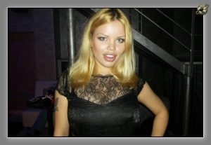 Blonde woman in black lace top beside a dim staircase with metal railings.