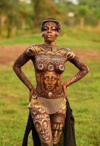 Performer with bronze and brown body paint in ornate patterns, hands on hips, grassy outdoor setting.