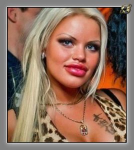 Close-up of a blonde woman with glossy lips and a pendant necklace, wearing a leopard-print top, dark crowd behind.