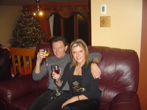 Two people on a burgundy leather sofa raising wine glasses, lit Christmas tree in background.