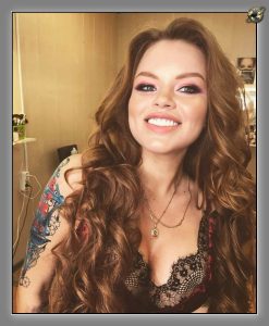 Smiling woman with long curly auburn hair, black lace bralette, layered pendant necklaces, and an arm tattoo, in warm light.