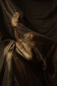 Figure with eyes closed beneath sheer dark gauze, draped across head and shoulders, in dim amber light.