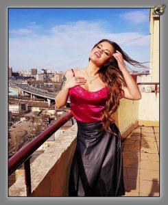 Woman with long hair blowing across her face, wearing a fuchsia satin camisole and black skirt on a rooftop balcony.
