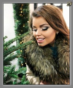 Woman in a coat with a thick grey fur collar, smiling downward beside evergreen branches strung with warm fairy lights.