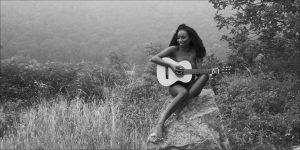 Black and white photo of a seated woman with a guitar on a rock in misty hills.