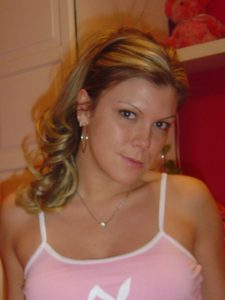 Woman leaning forward on a striped bed in a pink top and white thong, arms braced, looking toward the camera.