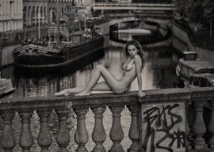 Black and white seated figure on stone railing above a canal with barges.