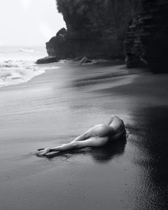 Black and white shot of a nude figure lying on wet sand beside a cliff and surf.