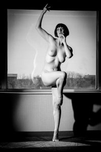 Black and white photo of a nude woman balancing on a window ledge, one arm raised, with a city view behind the glass.