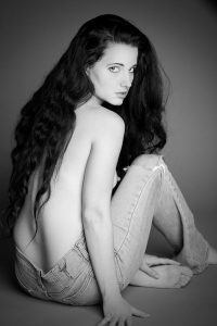 Black and white studio portrait of a woman with long wavy hair, seated on grey backdrop in jeans, looking over her shoulder.