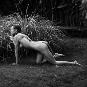Black and white photo of a nude figure crouched low on lawn grass, tall reed clumps and dense foliage behind.