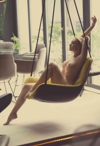 Nude woman in yellow chair hung on ropes, arm raised, glass wall and plants.