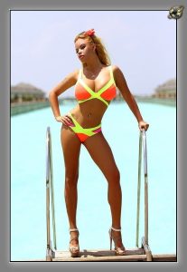 Woman in orange and yellow bikini posing on a pool ladder with pale blue water and resort buildings behind her.