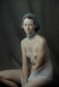 Seated nude model in pearl headpiece and choker, sheer white drape over lap, muted olive backdrop.