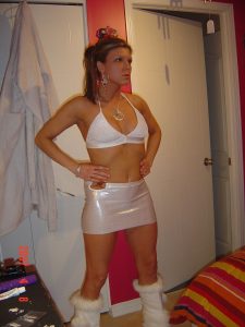 Woman kneeling on a red and orange striped bedspread in a pink bra top and matching white thong, red wall behind.