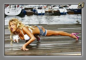 Woman in denim shorts and pink heels in a plank pose on a weathered wooden dock, boats moored behind.