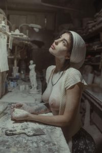 Woman in a cream knit top and headwrap kneads clay at a dusty counter, looking up among plaster sculptures.
