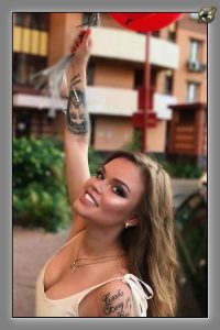 Smiling woman with arm tattoos beside a red umbrella, city building behind.