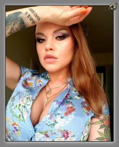Woman with raised arm, floral blouse, smoky eye makeup and forearm tattoo.