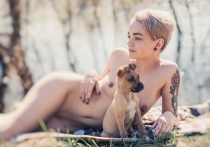 Short-haired nude model reclining outdoors on a blanket beside a small brown puppy, with blurred trees behind.