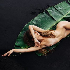 Overhead view of a long-haired woman lying on a green banana leaf over black sand.