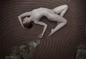 Nude figure lying on a sand surface with curved ripple patterns, pale skin against dark brown tones, grey stones at lower left.