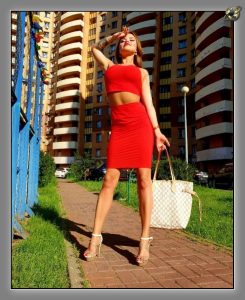 Woman in red two-piece dress and heels on brick path, cream handbag, sunlit towers behind.