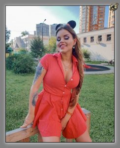 Auburn-haired woman in a red button-front playsuit seated on a bench on grass outdoors.