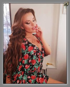 Woman with long curled hair in a black dress with red roses, one hand raised near her face, in a bright interior.