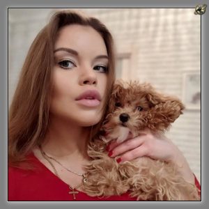 Woman in a red top with a gold cross necklace holding a curly apricot toy poodle against a pale background.