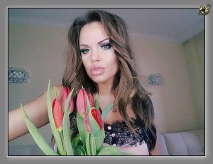 Woman with long wavy brown hair holding a bunch of red tulips near the camera, wearing a black lace top against a pale wall.