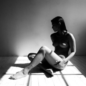 Black and white photo of a nude woman seated on a wooden floor, bright window light striping the boards and wall.