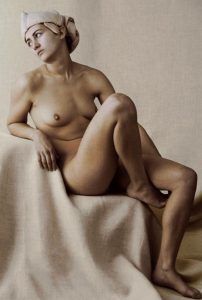 Nude model seated on pale linen, head wrapped in cream cloth, one knee raised, gazing sideways in warm light.