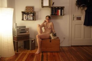 Nude woman wearing glasses sits on a wooden chest beside a glowing paper lamp and shelves.