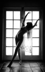 Black and white silhouette of a figure arching beside a tall paned window on tiled floor.