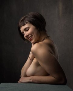 Short-haired nude model smiles with eyes closed, arms folded on a green table, dark grey backdrop.