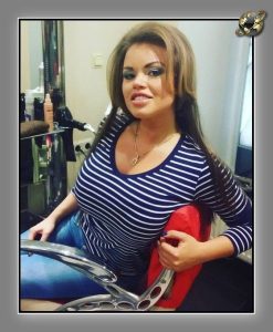 Woman with long brown hair in a navy striped top, seated in a salon chair before a mirror, red cape visible.