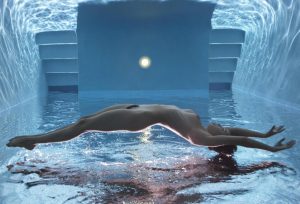 Nude figure floating horizontally underwater in a blue-tiled pool, a glowing light above the surface and steps behind.