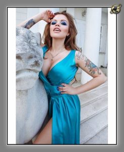 Auburn-haired woman in a teal satin wrap dress with a high slit, leaning on a stone lion sculpture outdoors.