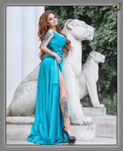 Woman in a long turquoise satin gown with a high slit, standing between white stone lion statues outdoors.