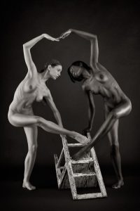 Black and white studio photo of two nude figures on a dark backdrop, one balancing a foot on a wooden step ladder, arms arched overhead.