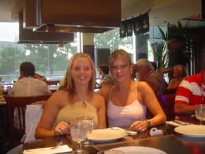 Two women in pale tops seated at a restaurant table, glasses and plates set, plants by window.