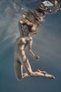 Nude figure suspended underwater with long hair floating upward, bent knees and arms, soft blue light filtering from the surface.