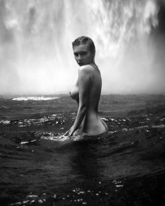 Black and white photo of a nude woman standing waist-deep in rippling water, looking over her shoulder at misty falls.